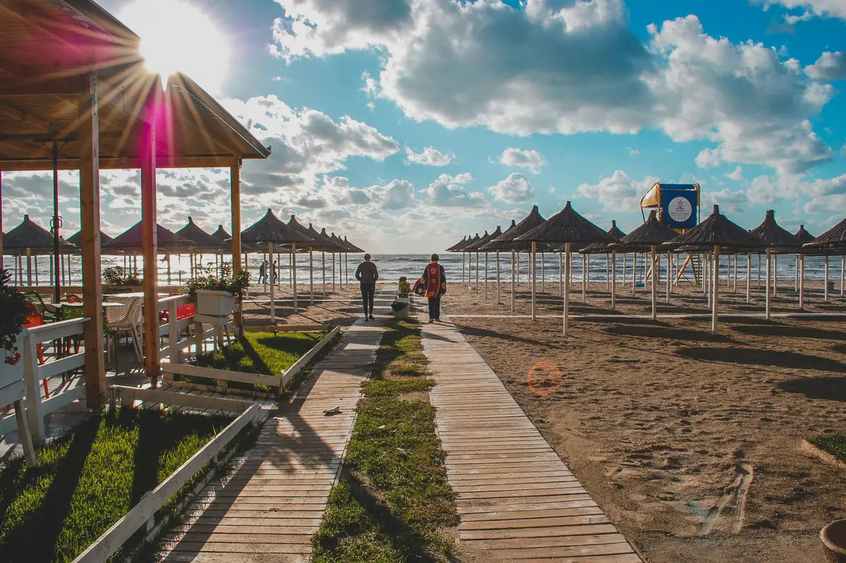 Une passerelle en bois traverse le sable entre des rangées de parasols de paille sur une plage de Durrës