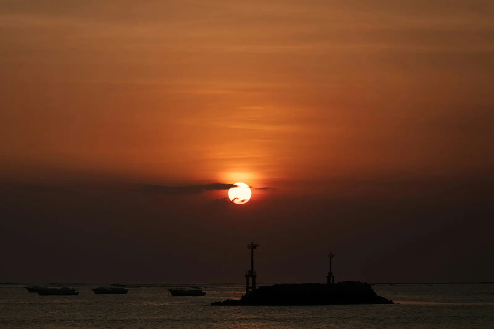Le soleil rouge se couche sur la baie de Jimbaran, au-dessus des bateaux de pêche et d’un îlot à balises en ombres chinoises