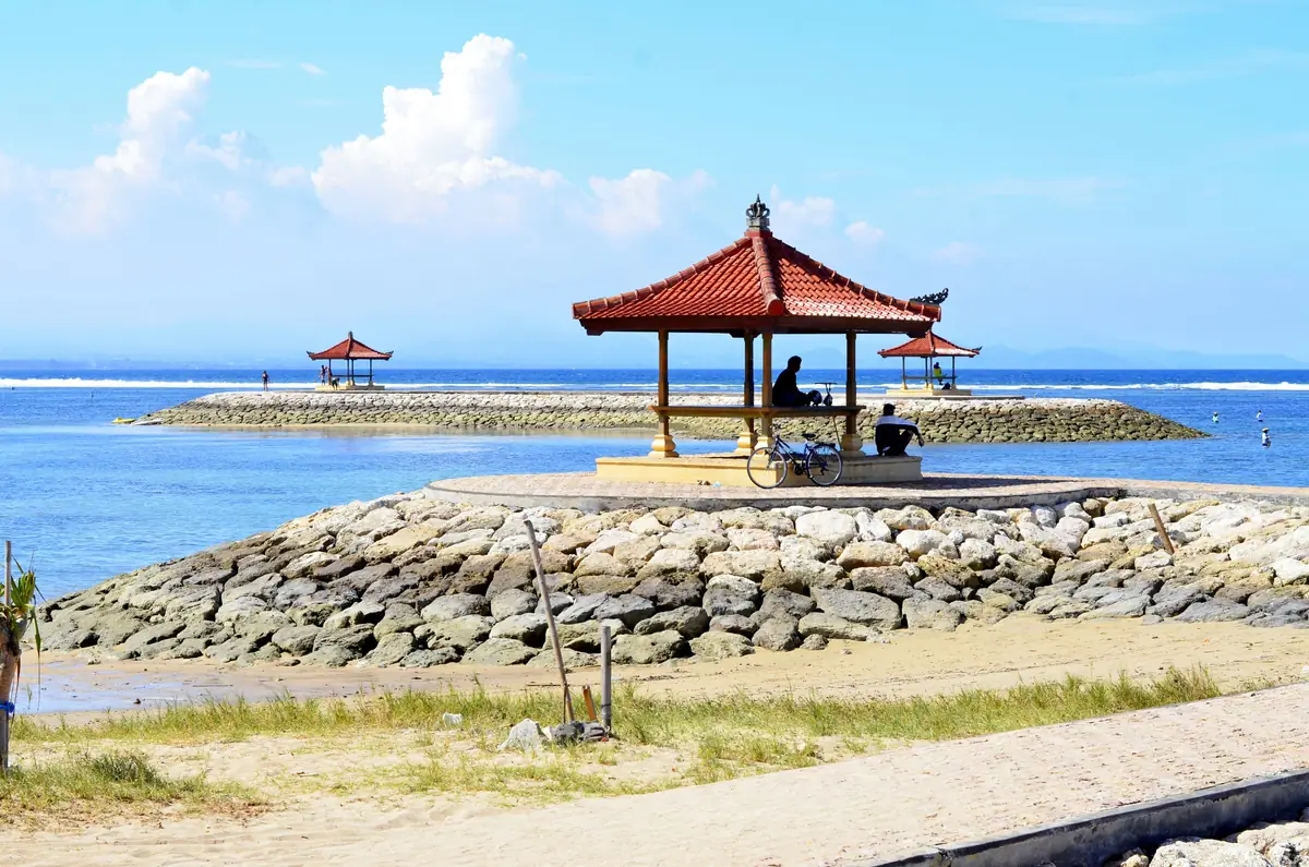 Un pavillon au toit de tuiles rouges au bout d’une digue de pierres, face au lagon bleu de Sanur protégé par le récif