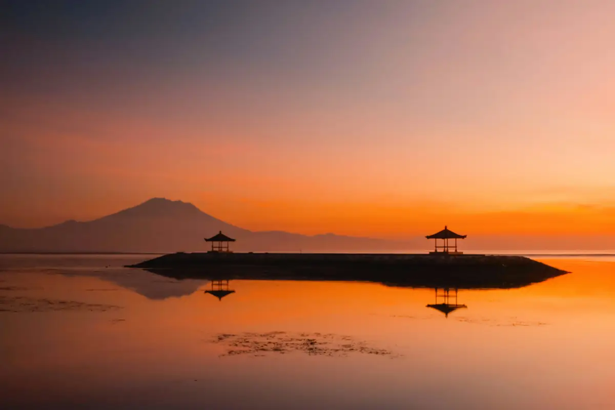 Deux pavillons balinais sur une digue de Sanur se reflètent dans le lagon à l’aube, devant la silhouette du mont Agung